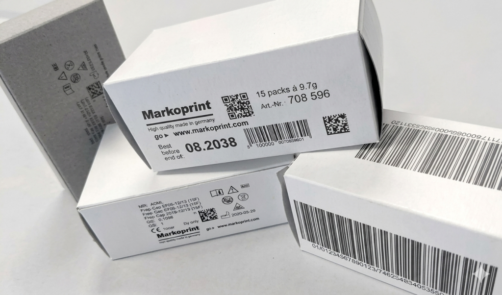 Stack of white cardboard boxes with Markoprint branding, barcodes, and QR codes visible on the labels.